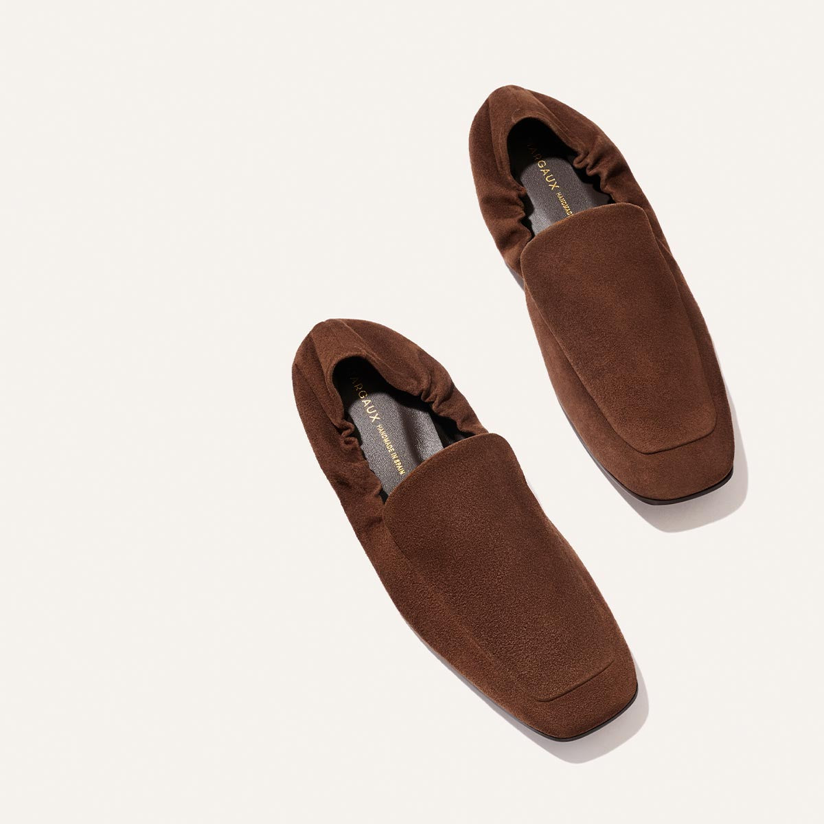 A pair of The Studio Loafer - Ganache Suede by Margaux, featuring brown suede in a minimalist design with square toes and elasticized heels, are placed on a light background.