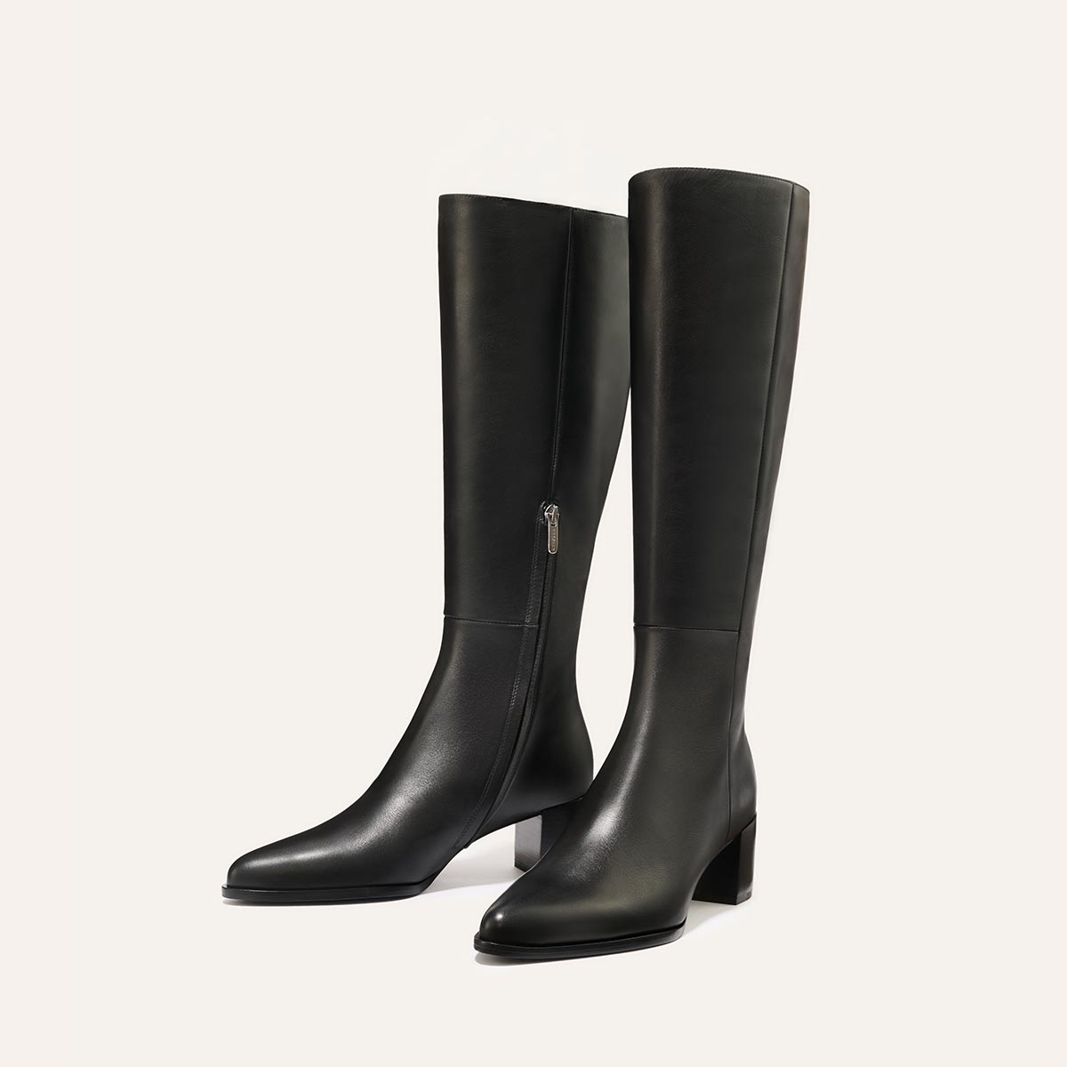 The Uptown Boot - Black Calf by margaux-development: sleek black knee-high leather boots with low block heels and inner side zippers, shown on a plain light background.