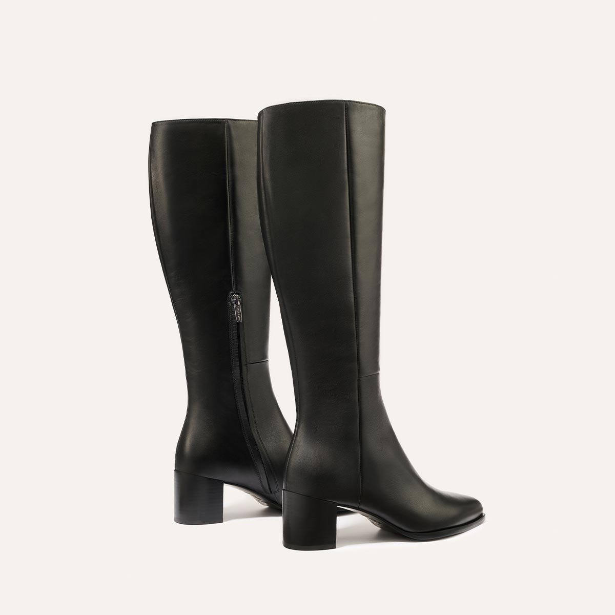 The Uptown Boot - Black Calf by margaux-development: two black leather knee-high boots with medium block heels and inner side zipper, shown against a plain light background—one in side profile, the other angled slightly backward.