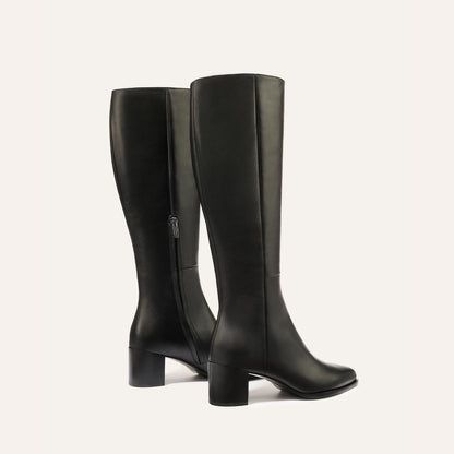 The Uptown Boot - Black Calf by margaux-development: two black leather knee-high boots with medium block heels and inner side zipper, shown against a plain light background—one in side profile, the other angled slightly backward.
