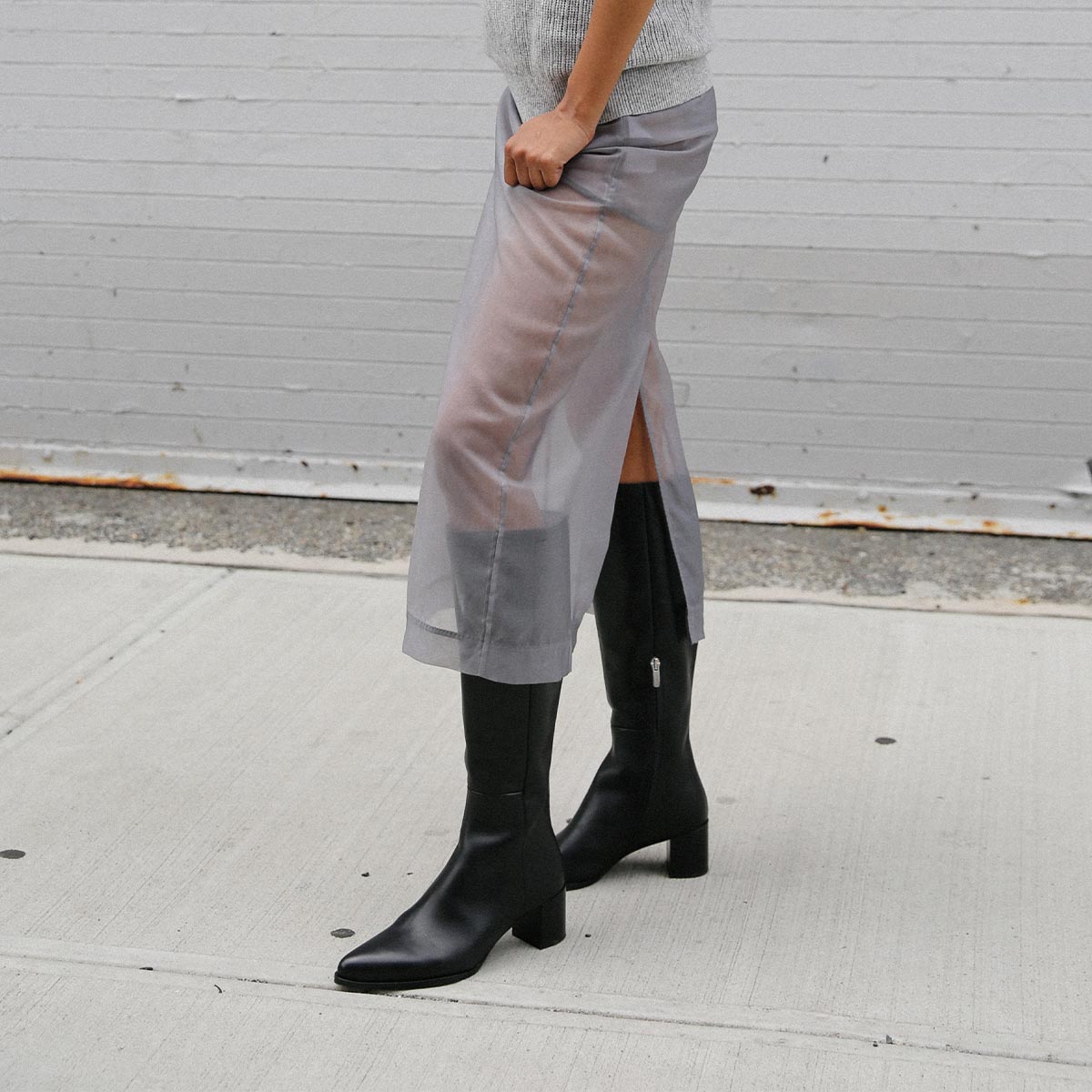 A person wearing a sheer gray midi skirt, light gray top, and margaux-development's The Uptown Boot in Black Calf stands on a sidewalk in front of a light gray brick wall.