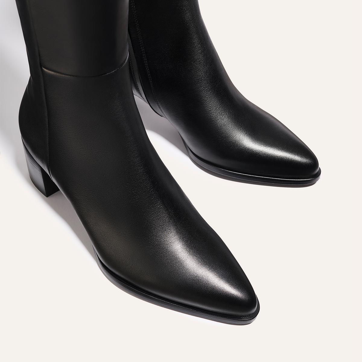 A close-up of The Uptown Boot - Black Calf by margaux-development, featuring black leather, pointed toes, and low block heels against a light background.