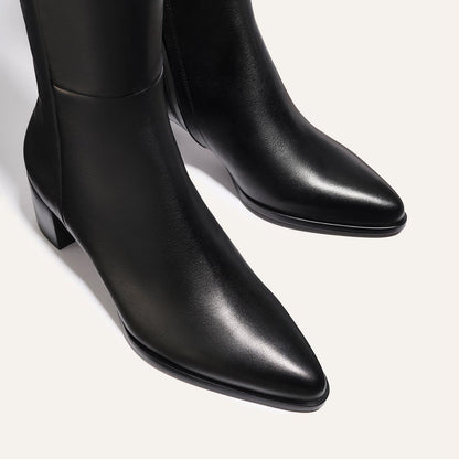 A close-up of The Uptown Boot - Black Calf by margaux-development, featuring black leather, pointed toes, and low block heels against a light background.