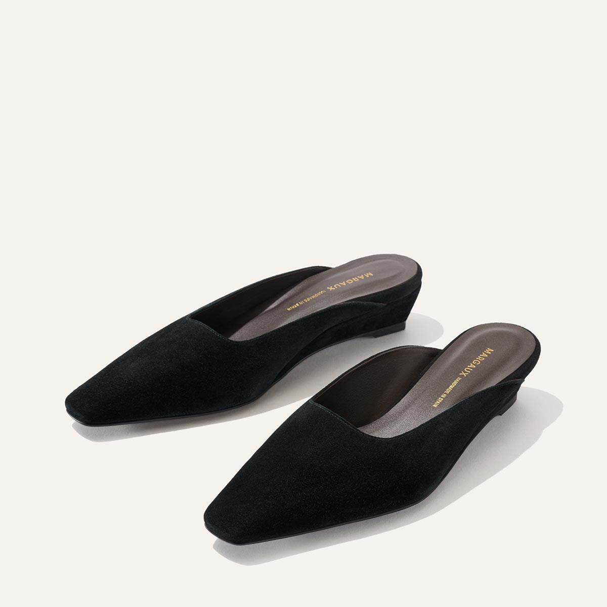 The Vesper Mule - Black Suede by Margaux, a pair of black suede slip-on wedge mules with pointed toes, is displayed against a plain, light background.