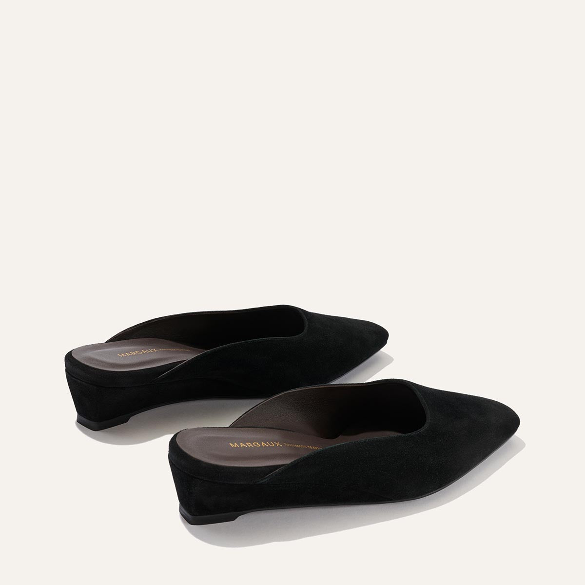 A pair of The Vesper Mule in black suede by Margaux, featuring a pointed toe, slip-on design, and low wedge heels, displayed on a light background.