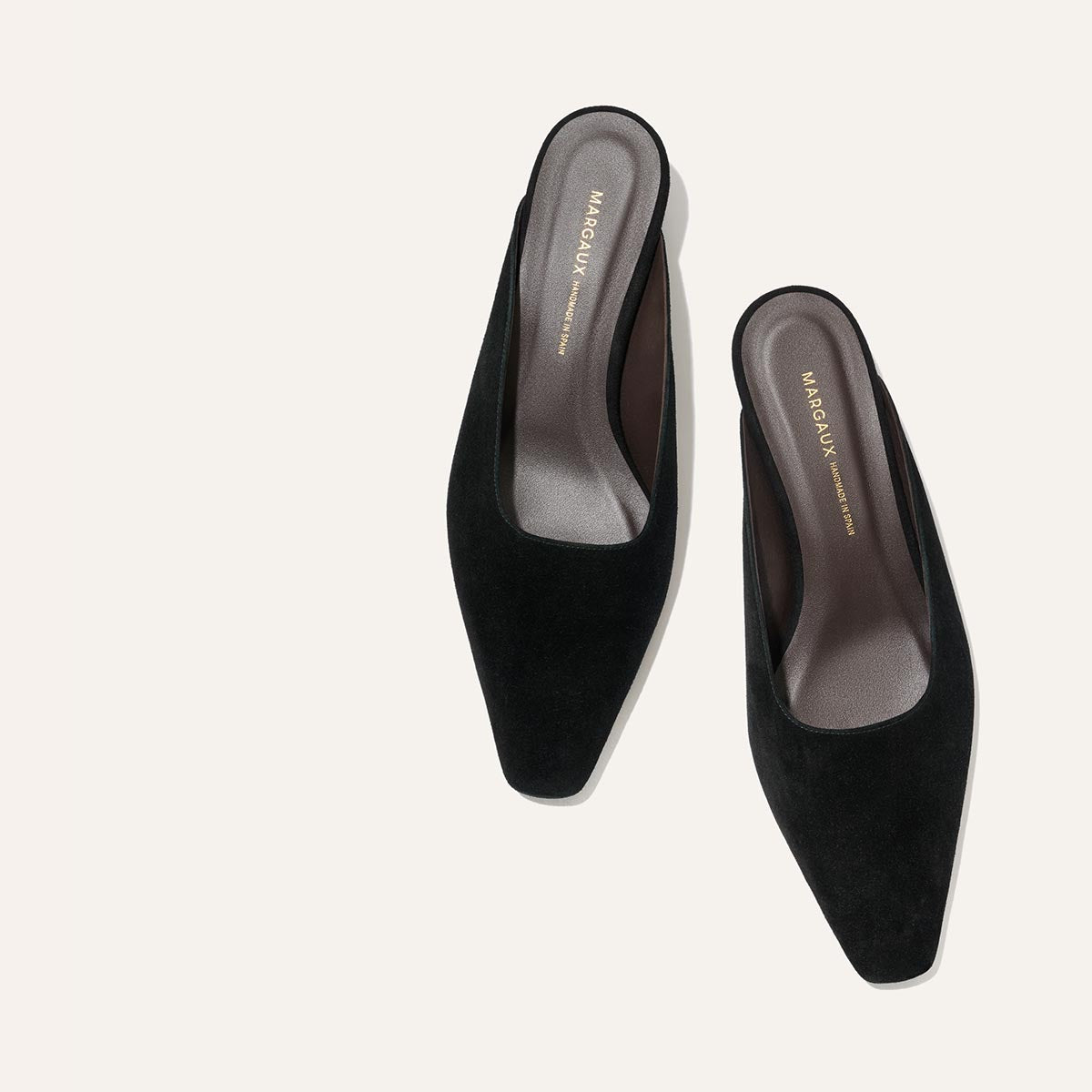 A pair of The Vesper Mule - Black Suede by Margaux, featuring a square toe and low heel in a slip-on design, is displayed on a plain light background.