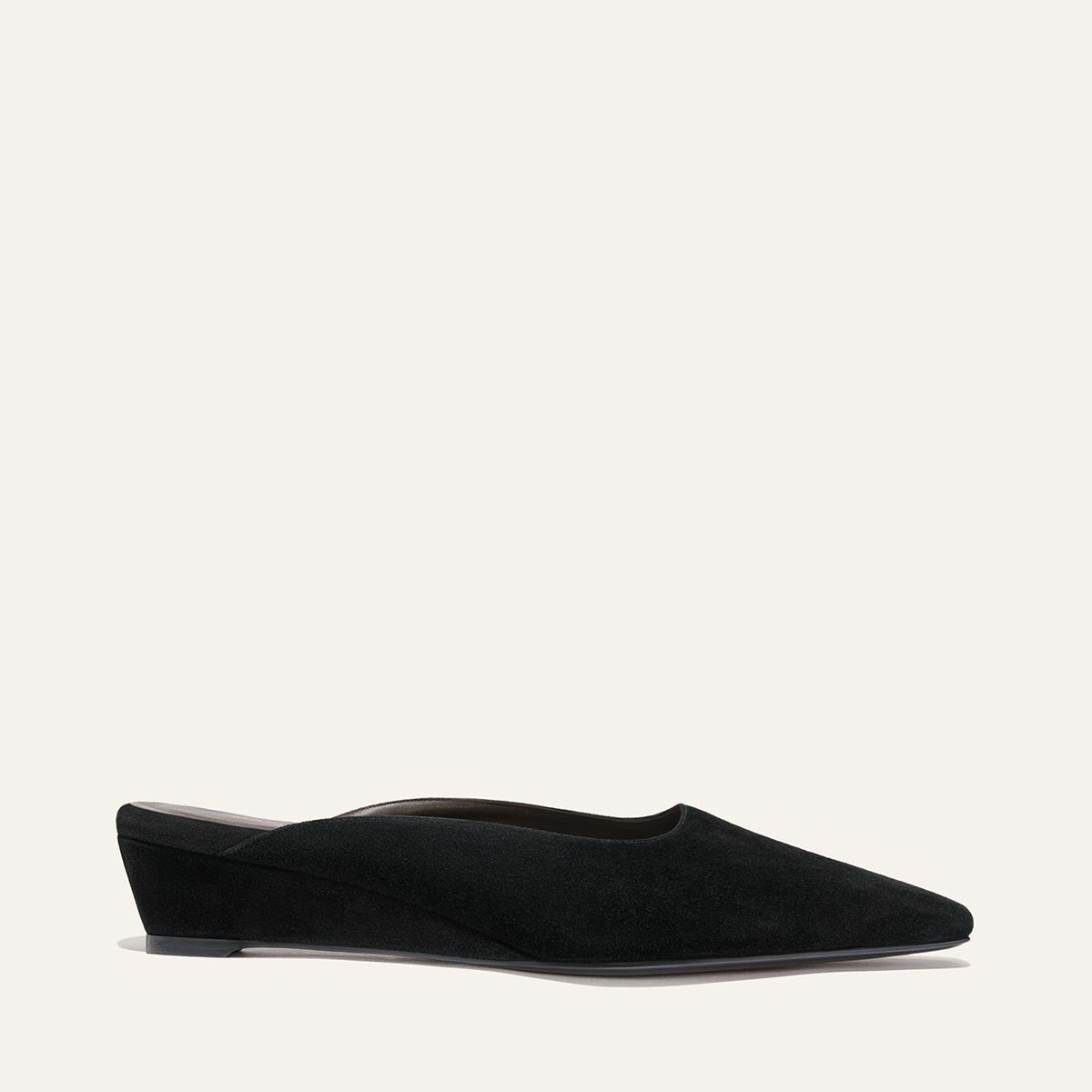The Vesper Mule by Margaux in black suede features a low wedge heel, pointed toe, and minimalist design, shown in a side view against a plain off-white background.