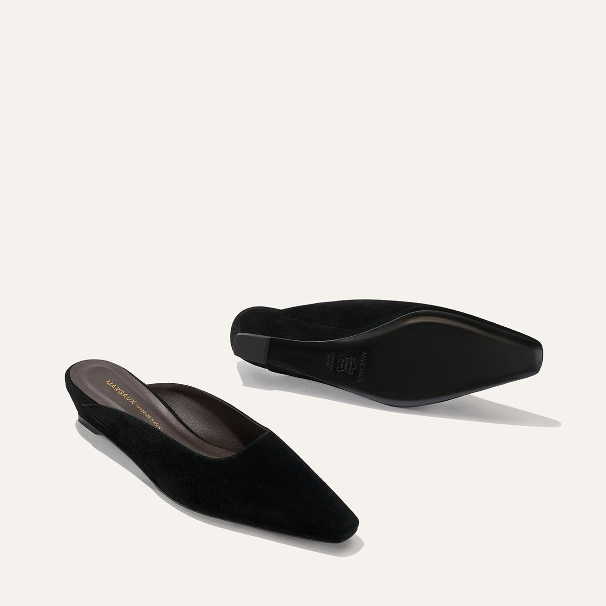 The Vesper Mule in Black Suede by Margaux is displayed on a light background, with one mule upright and the other flipped to reveal its sole.