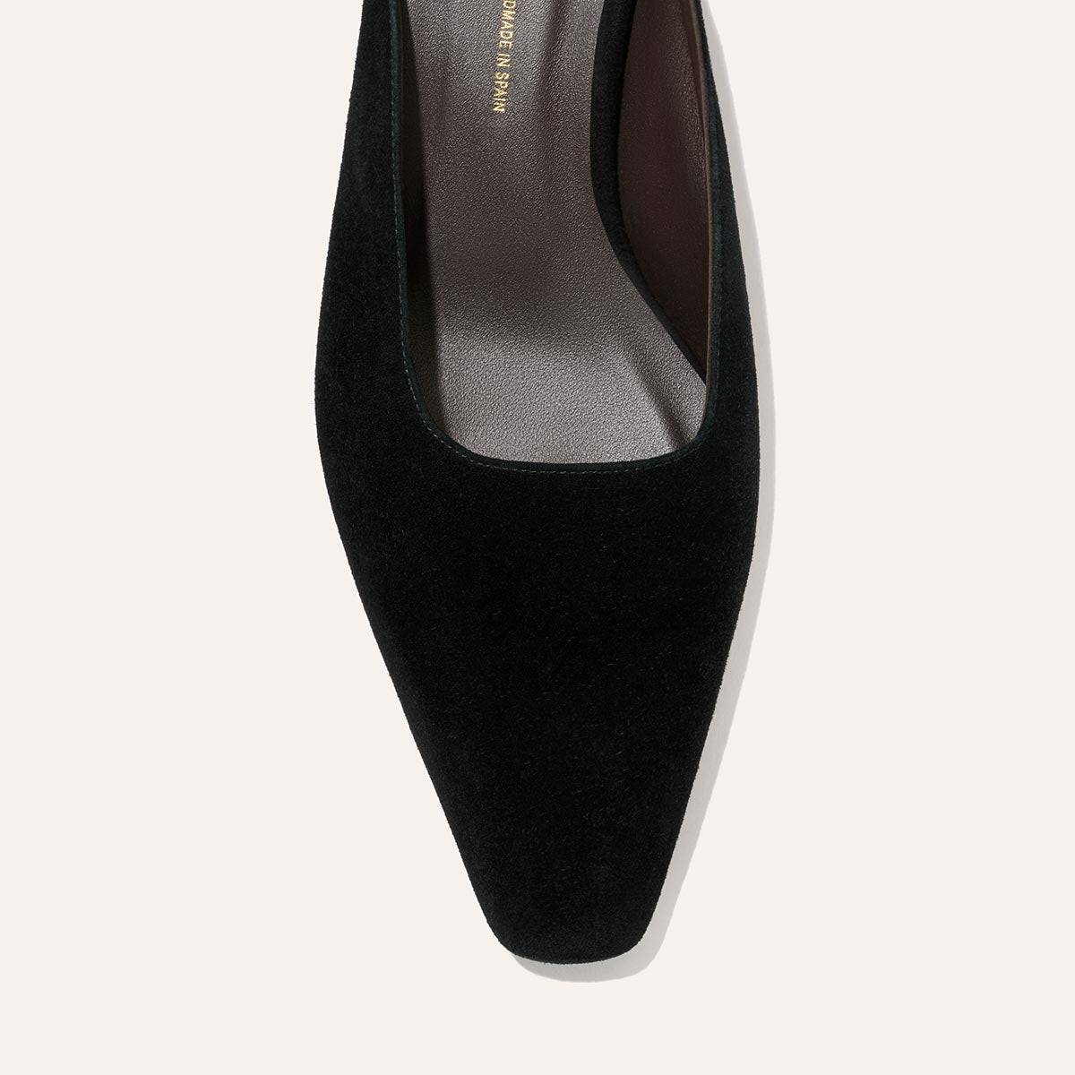A close-up of The Vesper Mule in black suede by Margaux, featuring a square toe on a light background, with part of the insole and upper material visible.