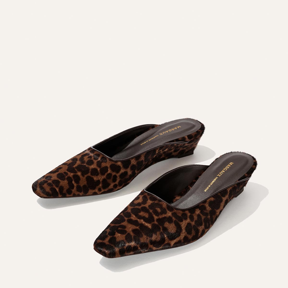 A pair of The Vesper Mule - Chocolate Leopard Haircalf by Margaux, featuring a brown and black leopard print, low wedge heels, and pointed toes, displayed on a plain white background.