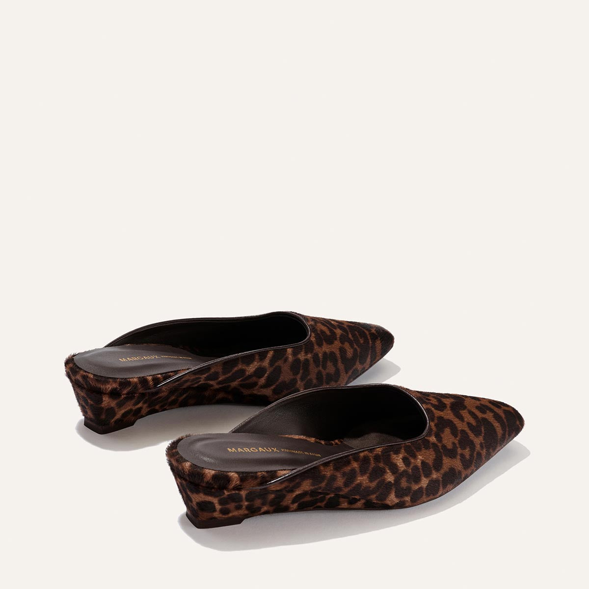 A pair of Margaux’s The Vesper Mule in Chocolate Leopard Haircalf, featuring pointed toes and wedge heels, is displayed from a slight angle against a plain light background.