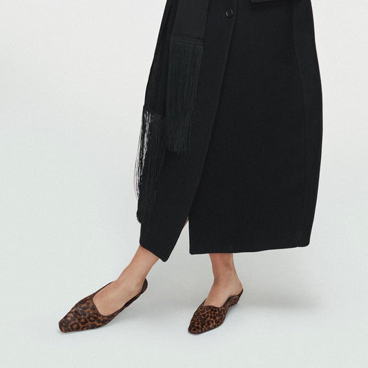 A person stands against a plain white background, wearing The Vesper Mule in Chocolate Leopard Haircalf by Margaux and a long black coat. The image is cropped to reveal only the lower half of the body.