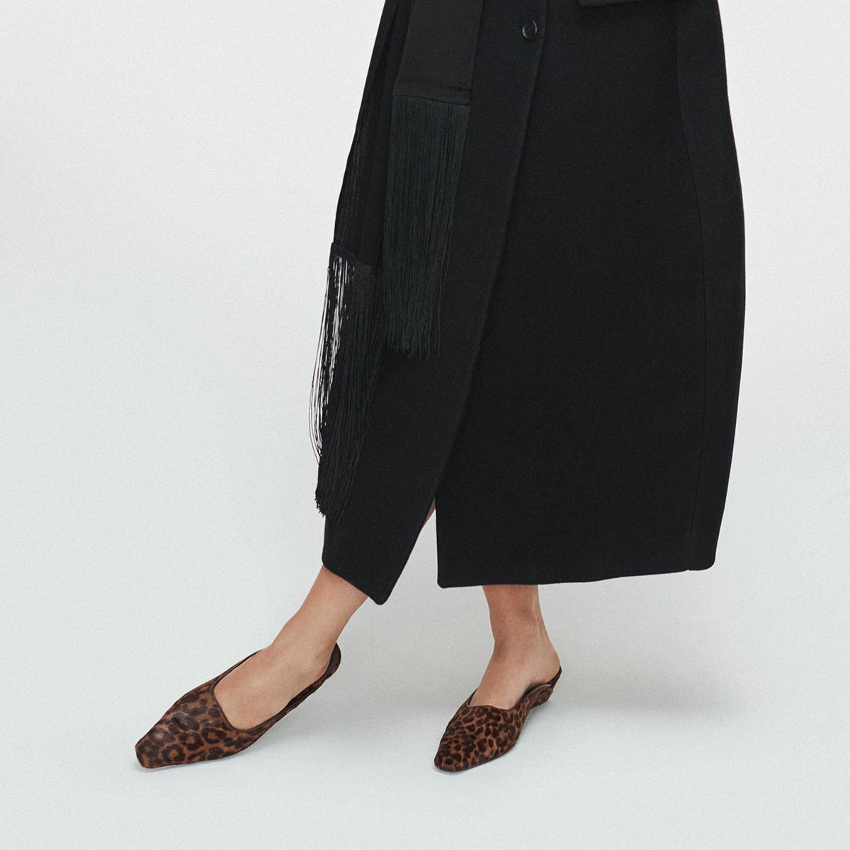 A person stands against a plain white background, wearing The Vesper Mule in Chocolate Leopard Haircalf by Margaux and a long black coat. The image is cropped to reveal only the lower half of the body.