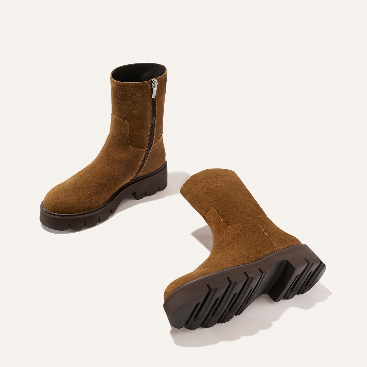 A pair of Margaux’s The Wells Boot in Cedar Suede—mid-calf brown boots featuring side zippers and chunky black rubber soles—are displayed on a white background. One boot stands upright, while the other lies on its side to showcase the tread.
