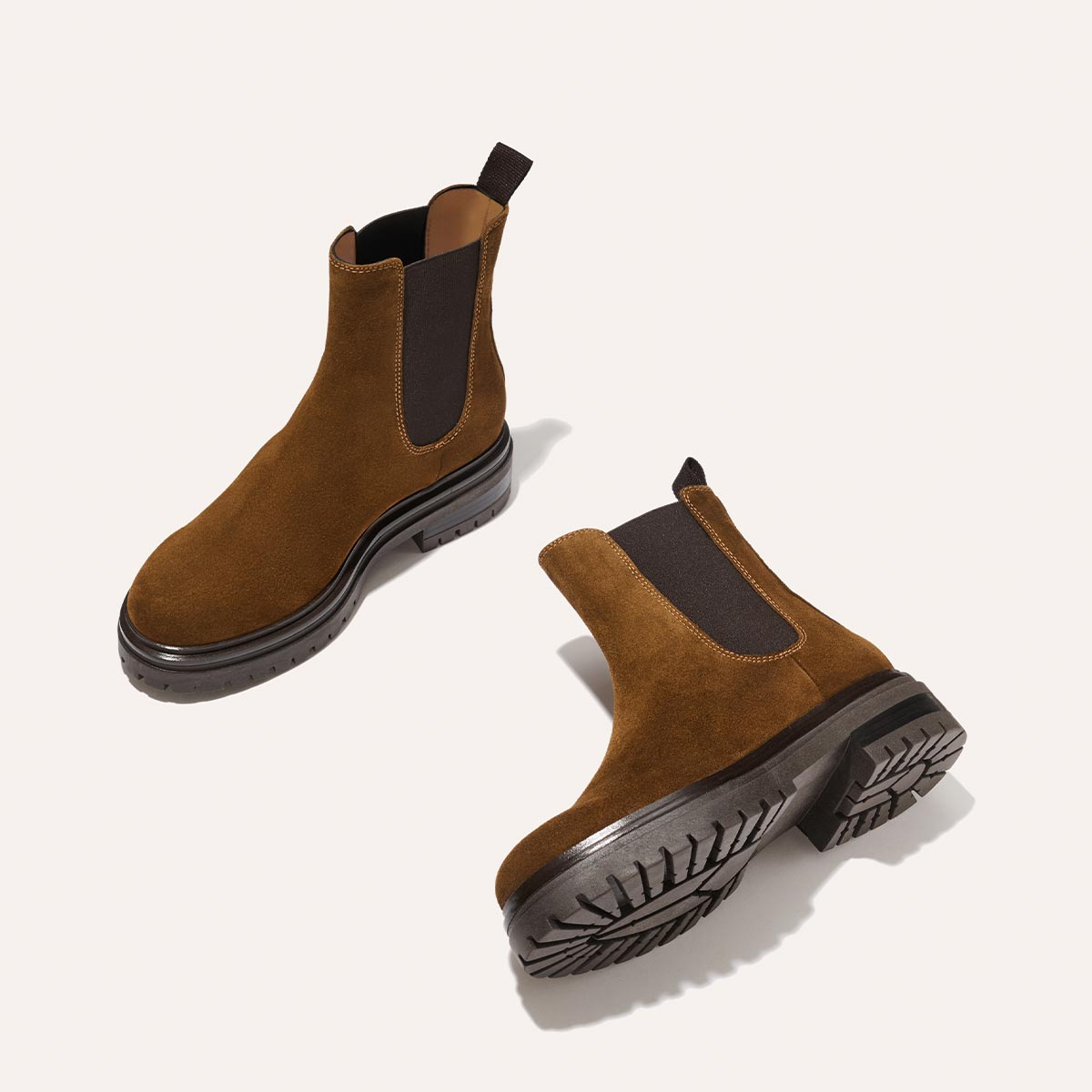 The West Chelsea Boot - Cedar Suede by Margaux: Italian brown suede boots featuring black elastic side panels and chunky lug soles, displayed on a light background with one upright and the other on its side to show the tread detail.