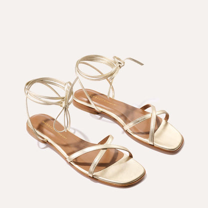 The Wrap Sandal - Champagne Nappa by Margaux features flat gold wrap design, ankle ties, and square toes, bringing getaway style to any look. Displayed on a neutral background.