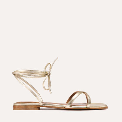 The Margaux Wrap Sandal in Champagne Nappa features metallic gold leather, two front straps, a slim ankle tie, and a small stacked heel—perfect for effortless summer style. Shown against a simple light background.
