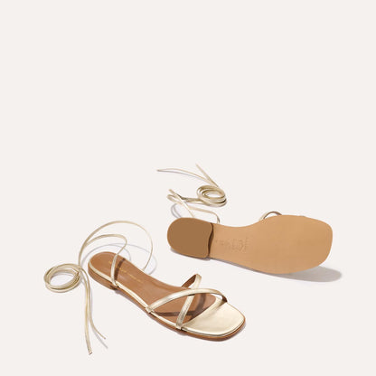 Two Margaux The Wrap Sandal - Champagne Nappa sandals are shown on a light background—one upright, one flipped to reveal the sole—highlighting their long ankle laces and summertime versatility, perfect for any getaway.