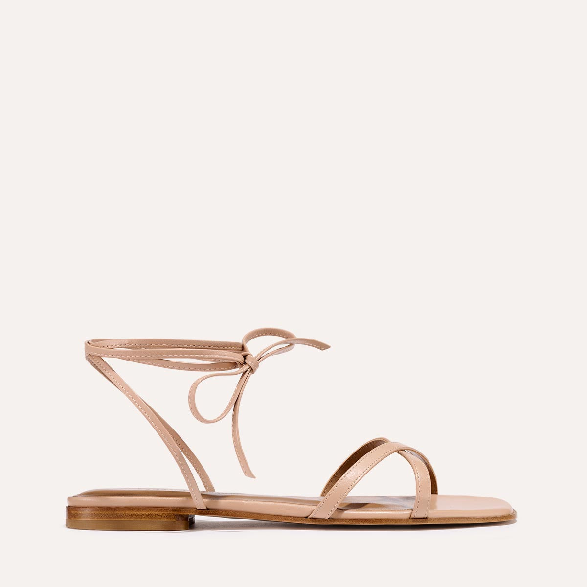 The Wrap Sandal - Rose Nappa by Margaux features thin straps, including one over the toes and another that wraps around the ankle with a bow tie. Its minimalist design and flat heel offer sophisticated style and summer versatility.