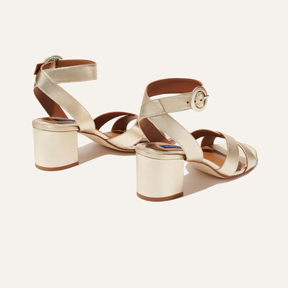 The City Sandal - Champagne Nappa by margaux-development: gold open-toe block heels with crisscross ankle straps, round buckles, and cushioned insoles for comfort, shown against a light background.