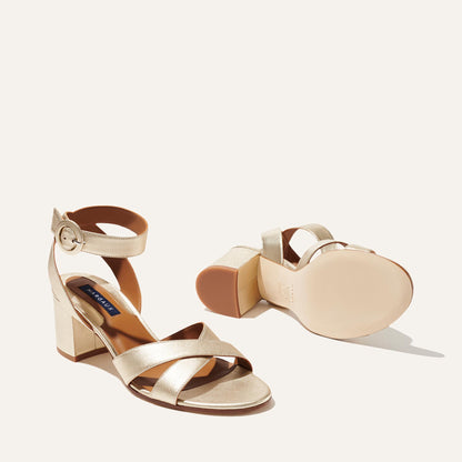 The City Sandal - Champagne Nappa by margaux-development features gold block heels, crisscross ankle straps with buckle closures, a cushioned insole, and a sturdy heel. One sandal stands upright; the other lies on its side.