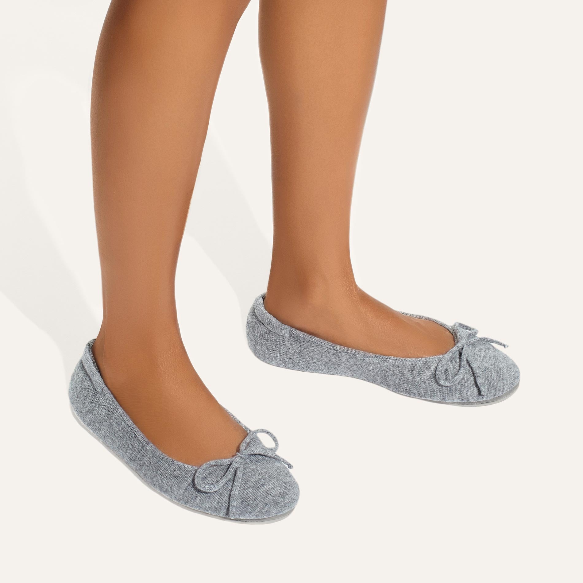 A person stands on a white background, wearing Margaux's The Slipper - Heather, light grey ballet flats with small bows and a plush quilted innersole for comfort. Only the lower legs and feet are visible, blending luxury and everyday style.