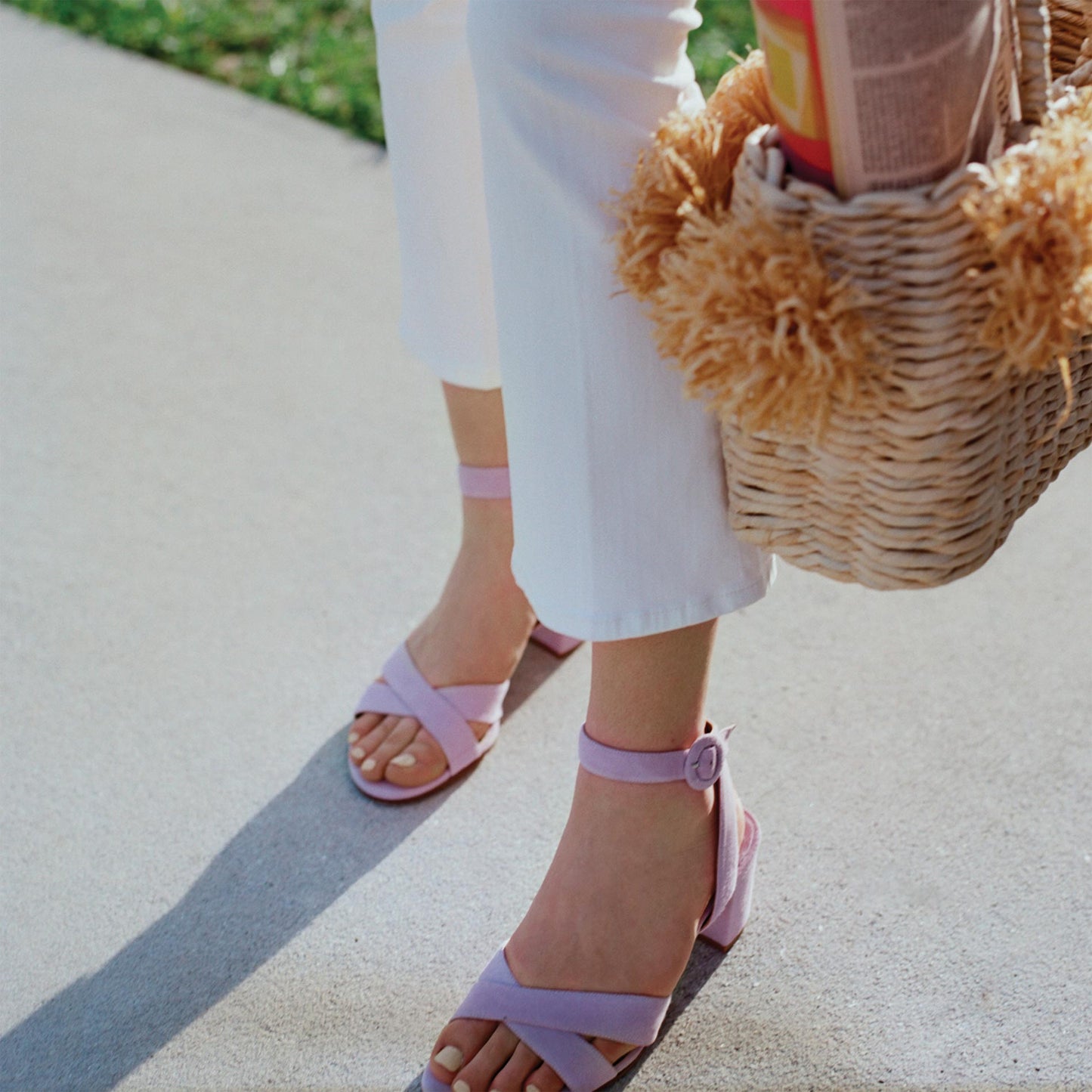 A person wearing margaux-development’s The City Sandal in Lilac Suede with a cushioned insole and white pants stands on a sidewalk, holding a woven straw bag with tan pom-poms. Grass shows in the background.