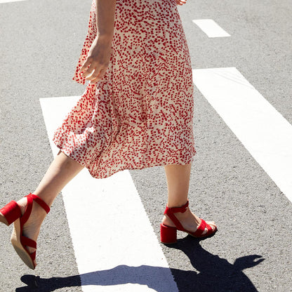 Wearing a white dress with red patterns and The City Sandal - Poppy Suede by margaux-development, she walks across a crosswalk on a sunny day, enjoying cushioned comfort with each stylish step.
