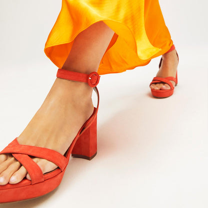 A person wears margaux-development’s The Platform Sandal in Chili Suede—red high-heeled sandals with ankle straps—paired with a flowing, bright yellow-orange dress. Only their lower legs and feet appear against a plain background.