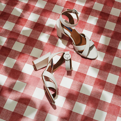 The Uptown Sandal - Ivory Nappa by margaux-development and a white wristwatch rest on a red and white checkered surface, sunlight creating soft shadows across the scene.