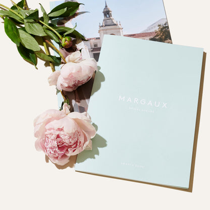 A pastel blue Bridal Swatch Kit from Margaux rests beside two pink peonies and a leafy stem, with a photo partially tucked beneath, evoking an exclusive bridal atelier experience.