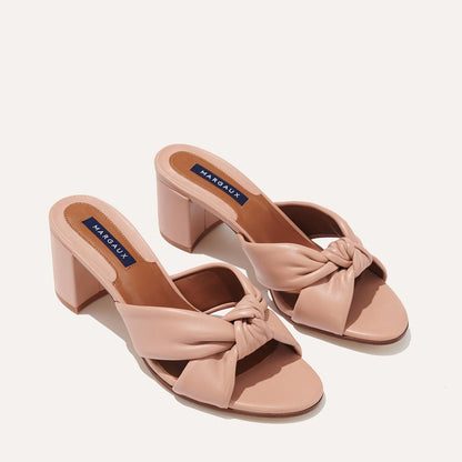 The Carmine Mule - Rose Nappa by margaux-development features blush pink, open-toe heeled sandals with plush knotted straps, a sturdy block heel, and a navy blue MARGAUX label on the insole, displayed against a plain light background.