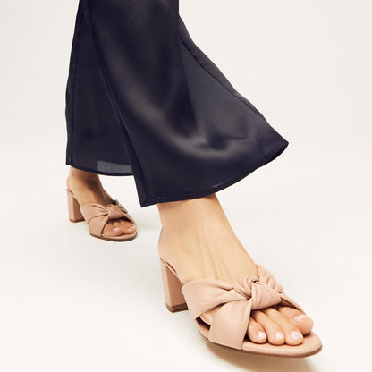 A person stands on a light background wearing The Carmine Mule - Rose Nappa by margaux-development, beige open-toe block heel sandals with plush knot detail, paired with black satin pants for a look of comfort and style.