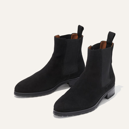 The Chelsea Boot - Black Suede by Margaux features elastic side panels, back pull tabs, and low block heels. This stylish pair is shown on a light beige background.