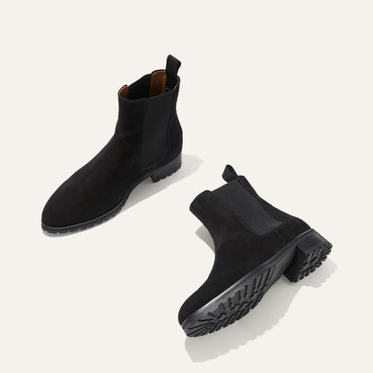 The Chelsea Boot - Black Suede by Margaux is shown on a light background. One boot stands upright while the other is tilted to display the textured sole, highlighting the elastic side panels and pull tabs.