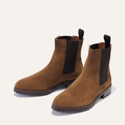 The Chelsea Boot - Moss Suede by Margaux features brown suede, black elastic side panels, dark soles, and pull tabs at the back, shown on a light background.