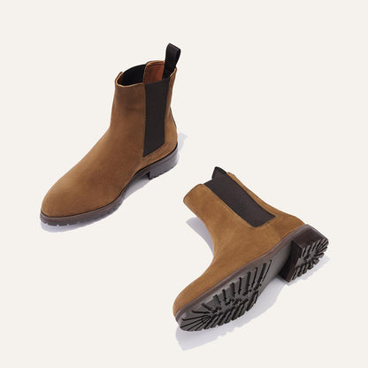 The Margaux Chelsea Boot in Moss Suede features black elastic side panels and pull tabs. One boot stands upright, while the other shows its ridged rubber sole on a light background.