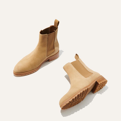 The Chelsea Boot - Sand Suede by Margaux: A pair of tan suede boots with brown elastic side panels, pull tabs, and chunky wooden soles are shown on a light background. One boot stands upright; the other lies on its side.