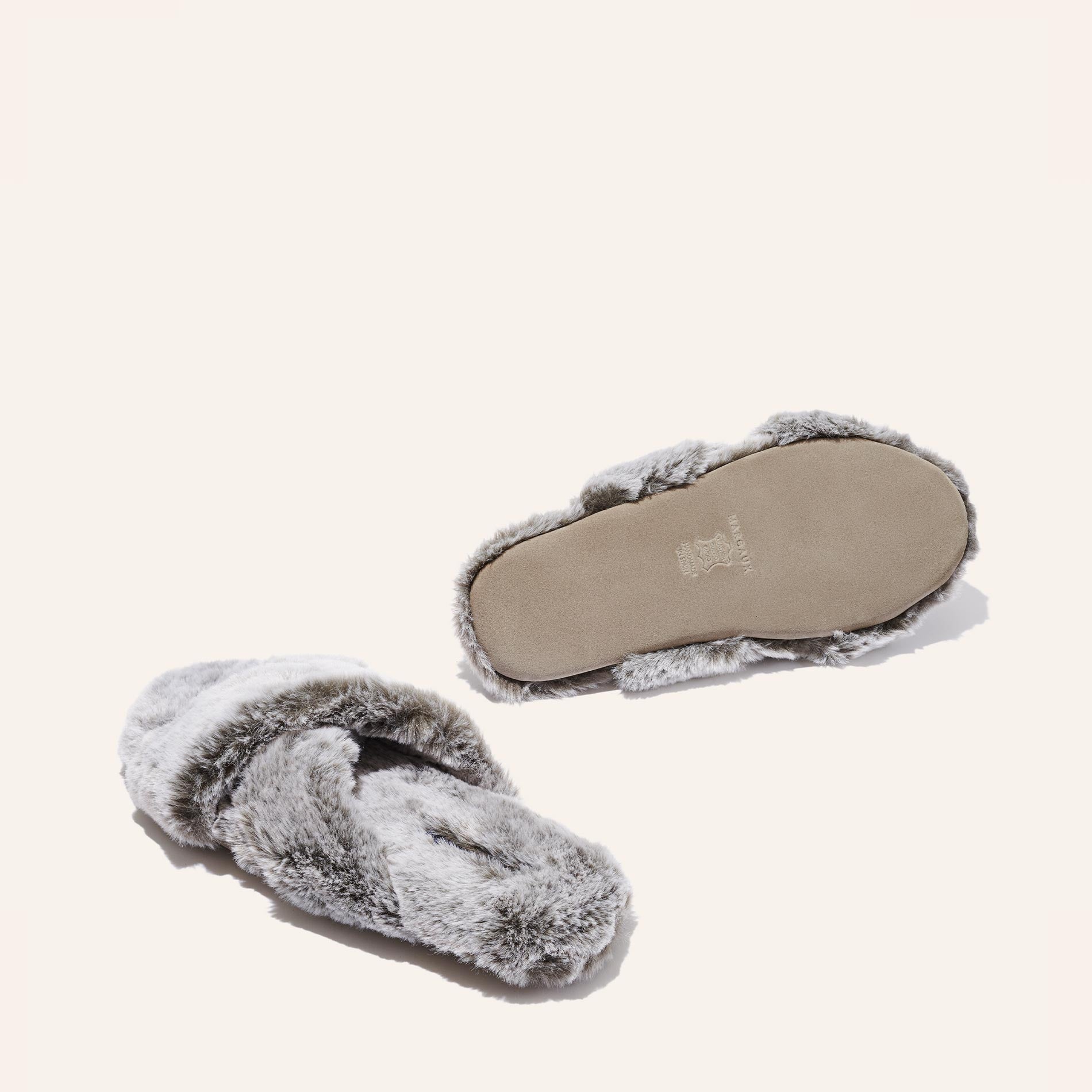 Margaux’s The Cozy Slipper - Grey, featuring a stylish x-strap design and plush faux fur, is displayed on a light background with one slipper upright and the other turned over to show the sole.