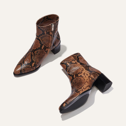 The Downtown Boot First Edition - Espresso Python Embossed by Margaux: brown, mid-heeled, western-style ankle boots with a snakeskin pattern and side zippers. One boot stands upright, the other lies on its side to reveal the sole.