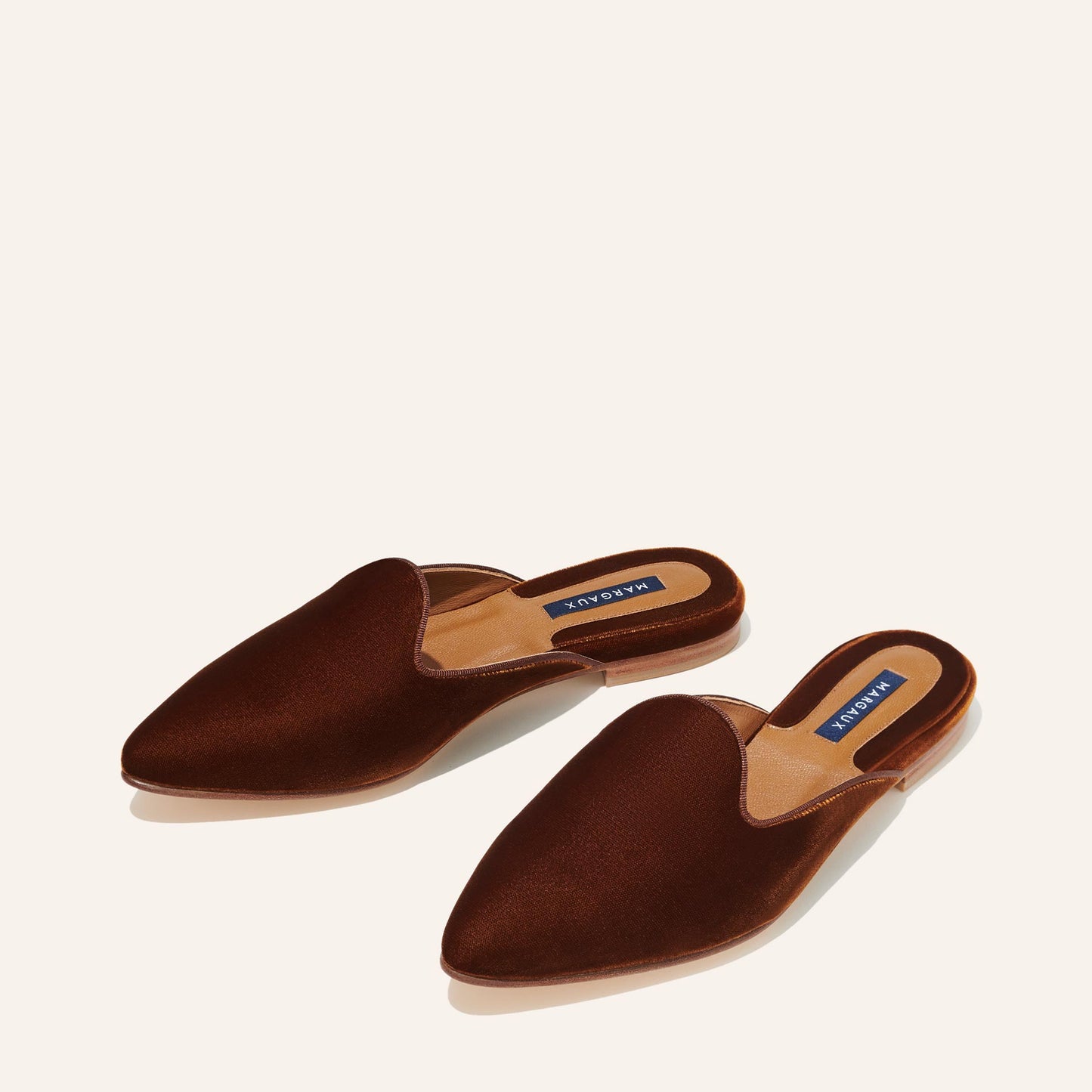 The Mule - Rust Velvet by Margaux: brown velvet mule slides with pointed toes and tan insoles, each adorned with a blue Margaux label—combining stylish design and comfortable wear.