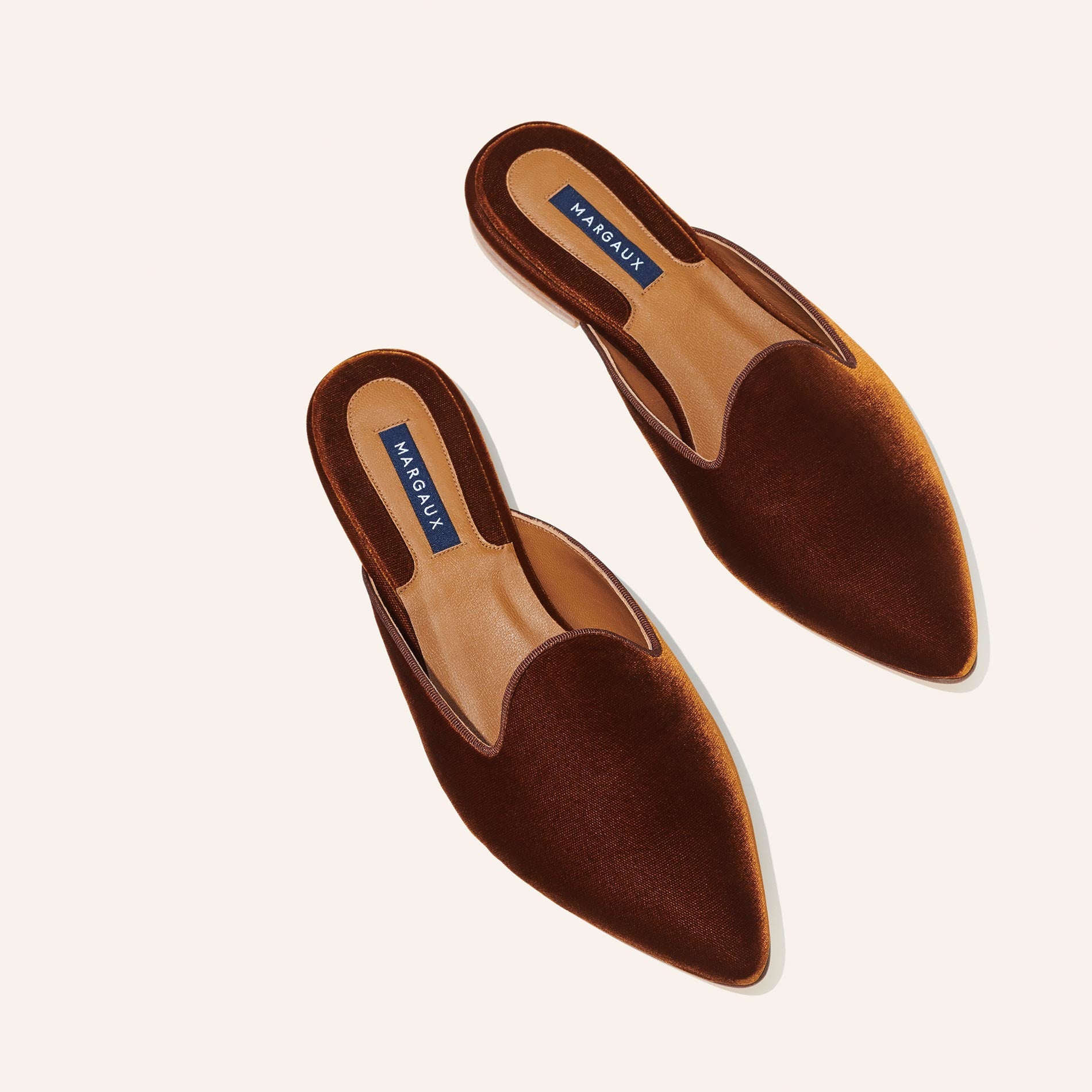 A pair of The Mule - Rust Velvet pointed-toe slide mules by Margaux, featuring tan insoles and brand labels, displayed on a plain light background.
