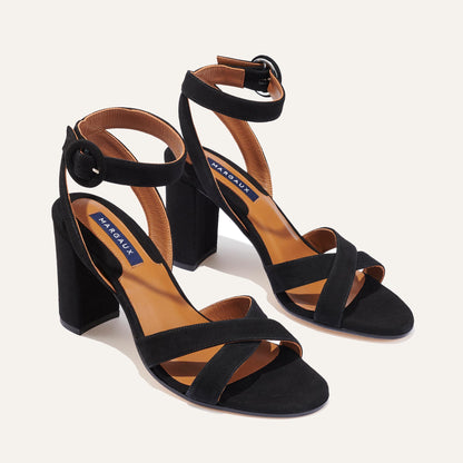 The Uptown Sandal - Black Suede by margaux-development features thick block heels, crisscross toe straps, round-buckle ankle straps, and a tan insole with a navy Margaux logo. Designed for all-day comfort.