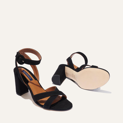 The Uptown Sandal - Black Suede by margaux-development features block heels, ankle straps with buckles, and a comfortable sole—one shoe stands upright while the other lies on its side, highlighting its design for all-day wear.