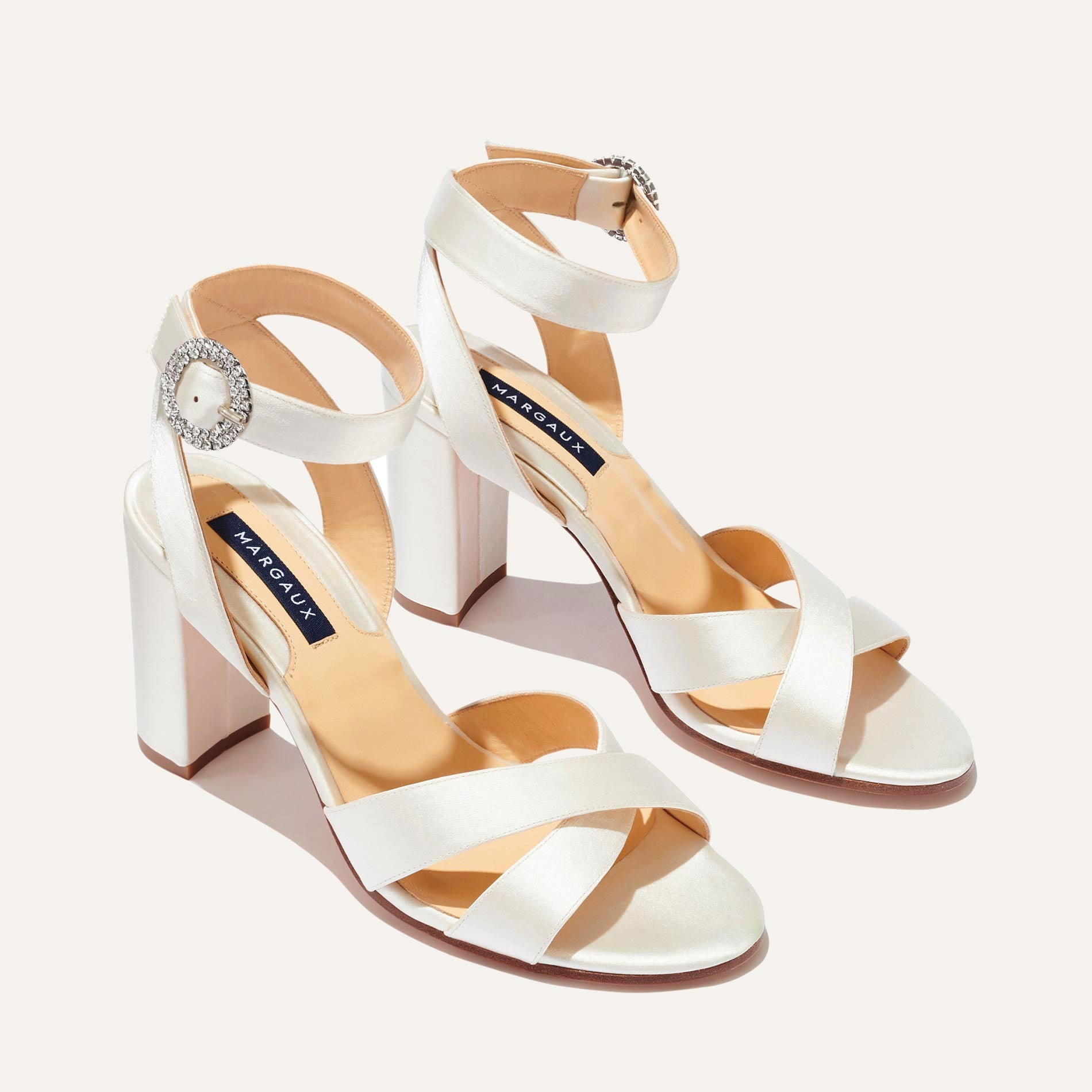 The Uptown Sandal - Ivory Satin with Crystal Buckle by margaux-development features crisscross straps, a chunky heel, and ankle strap with a crystal buckle—elegant formal sandals perfect for any occasion.