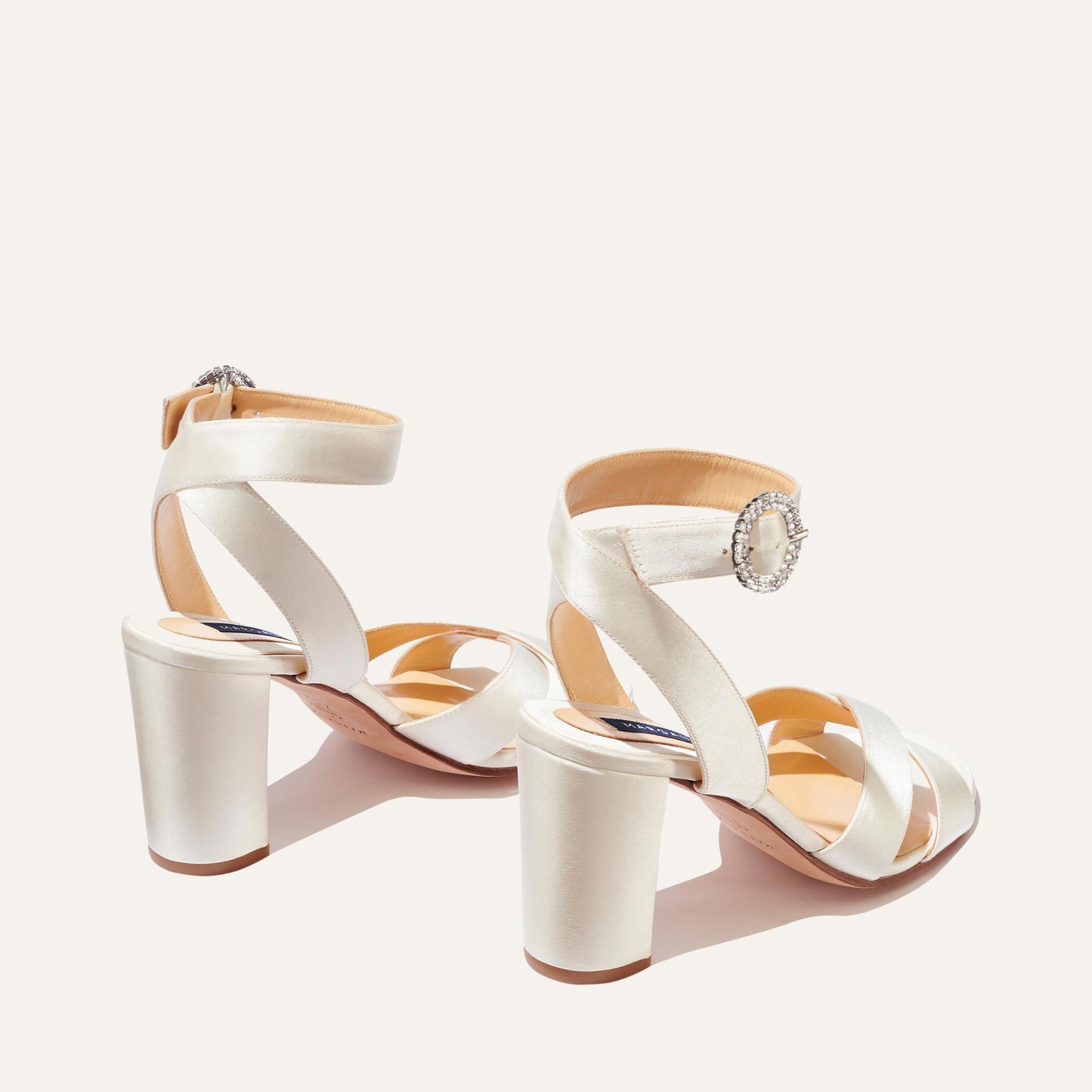 A pair of The Uptown Sandal - Ivory Satin with Crystal Buckle by margaux-development, featuring crisscross and ankle straps, shown on a light background—ideal formal sandals for any special occasion.