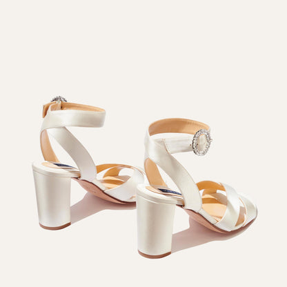 A pair of The Uptown Sandal - Ivory Satin with Crystal Buckle by margaux-development, featuring crisscross and ankle straps, shown on a light background—ideal formal sandals for any special occasion.