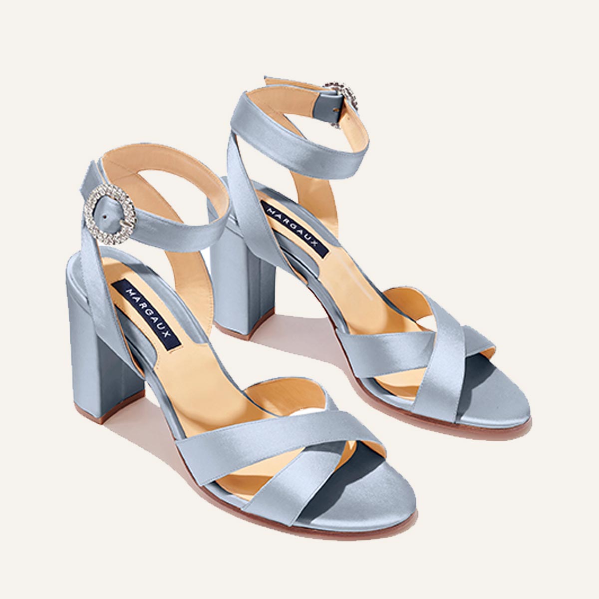 The Uptown Sandal by margaux-development features powder satin, an open toe, block heel, crisscross and ankle straps with a crystal buckle, cushioned insoles for comfort, and a blue Margaux label on the beige insole.